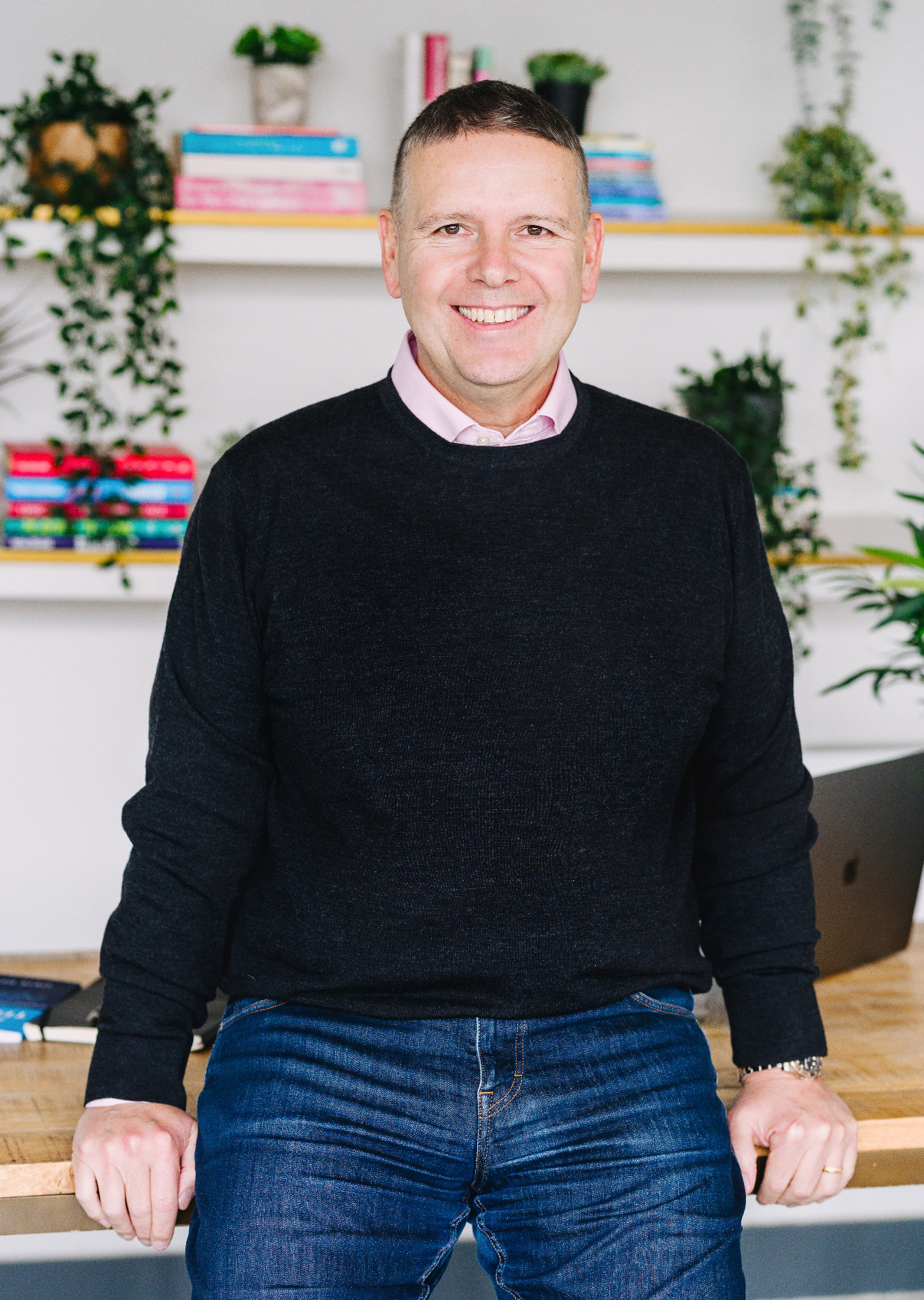 Steve Matthews smiling, leaning against his desk in a sunlit studio